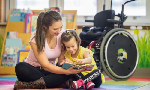 A woman sitting on the floor reading with a child and there is a wheelchair behind them.