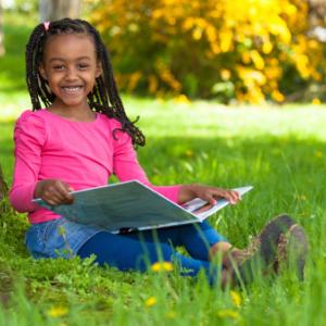 A girl sitting in the grass with a book and smiling at the camera.