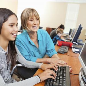 A smiling woman sitting next to a smiling girl who is working on the computer.