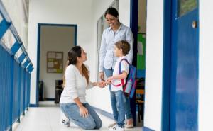 A woman on her knees talking to a boy and another woman in a school hallway.