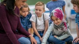 four children sitting on the floor, one girl in a hijab, listening to a woman